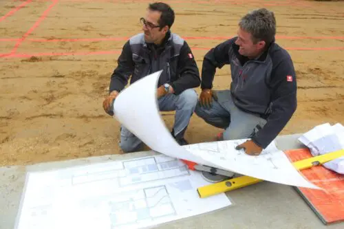 Zwei Bauwerk-Mitarbeiter hocken auf der Baustelle und besprechen ausgebreitete Baupläne