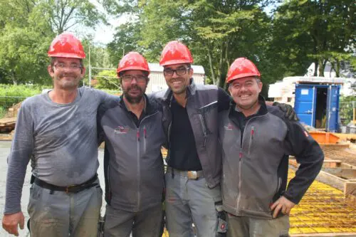 Vier Bauwerk-Mitarbeiter mit roten Schutzhelmen stehen lächelnd Arm in Arm auf einer Baustelle