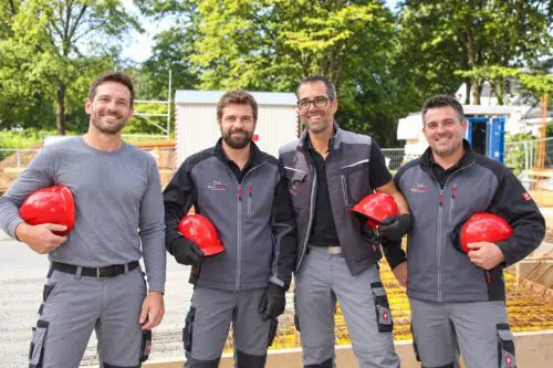 Vier lächelnde Bauwerk-Mitarbeiter in Arbeitskleidung stehen nebeneinander auf einer Baustelle und halten rote Schutzhelme in der Hand